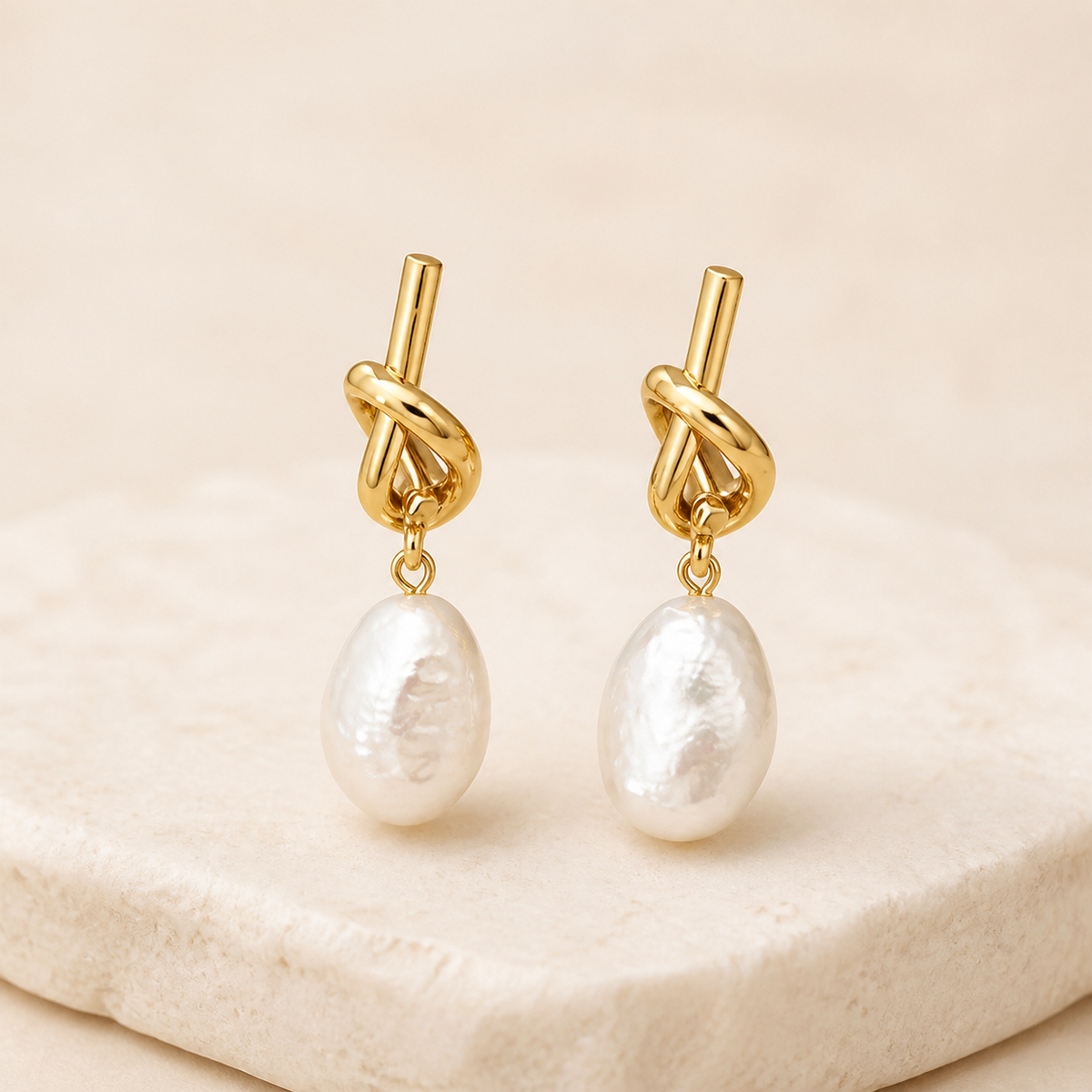 Gold sculptural knot earrings with white baroque pearl drops on an ivory studio backdrop