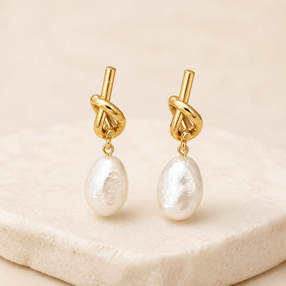 Gold sculptural knot earrings with white baroque pearl drops on an ivory studio backdrop